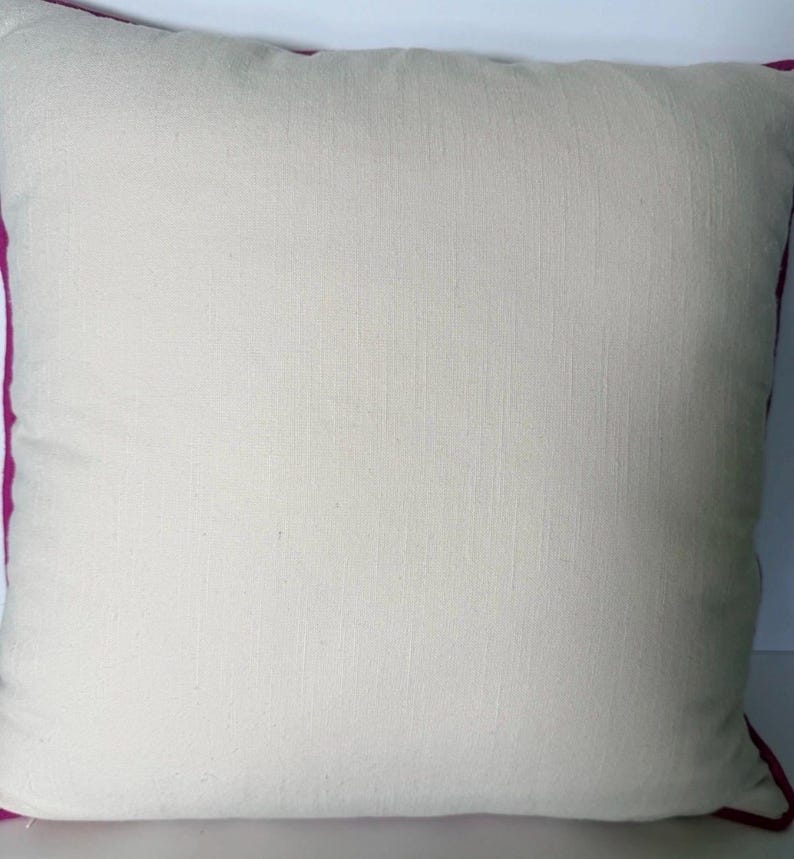 May include: Square decorative pillow with a cream-colored center and a vibrant magenta border. The pillow has a textured appearance, suggesting a natural fabric like linen or cotton. The design is simple and elegant, suitable for various home decor styles.