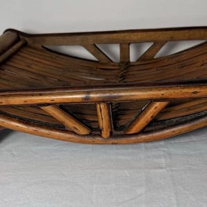 May include: A rectangular brown bamboo tray featuring a curved, slatted base and a raised frame with angled support struts. The tray is supported by four short, sturdy legs and displays a natural wood grain finish with dark brown accents throughout.