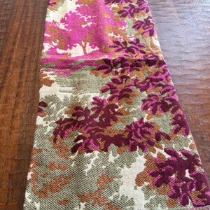 May include: A rectangular scarf with a colorful floral pattern. The scarf features a design of trees and foliage in shades of pink, burgundy, green, and brown. The scarf has a fringed edge and is displayed on a wooden surface.