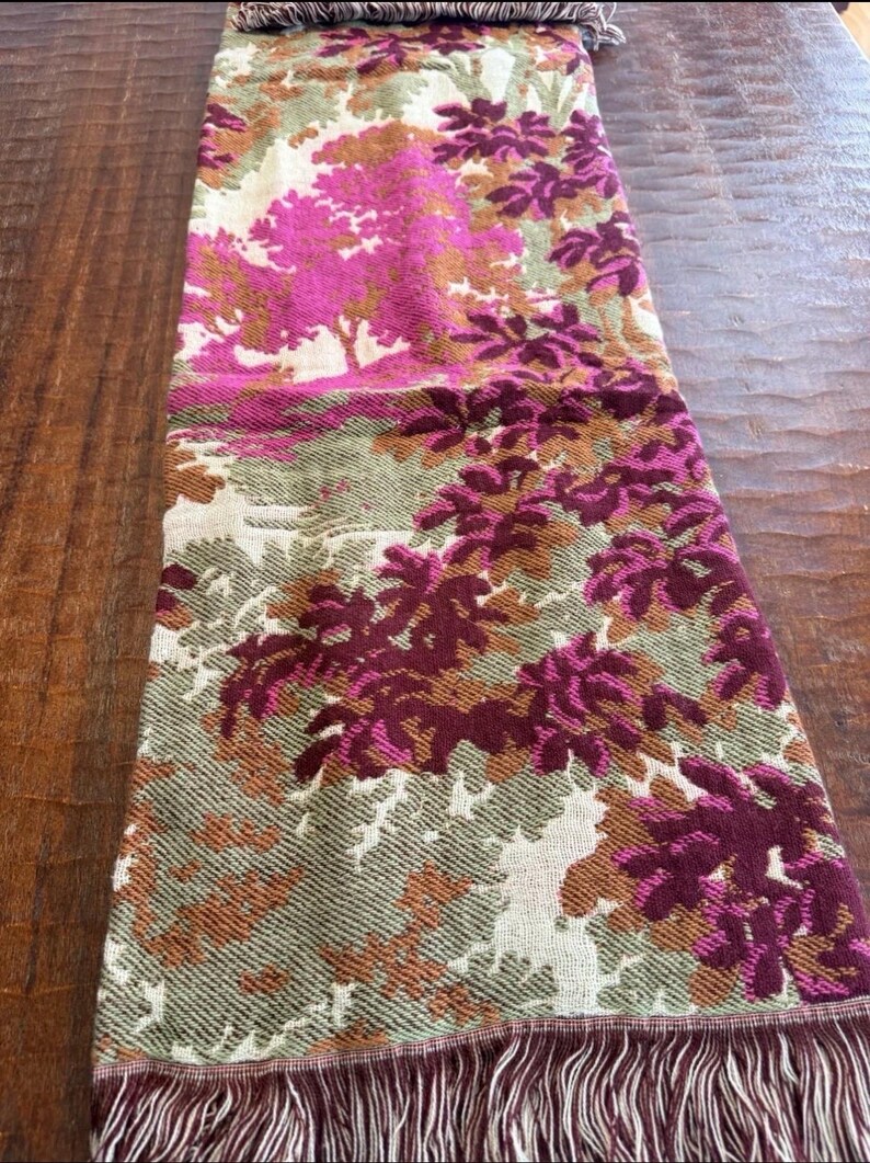 May include: A rectangular scarf with a colorful floral pattern. The scarf features a design of trees and foliage in shades of pink, burgundy, green, and brown. The scarf has a fringed edge and is displayed on a wooden surface.
