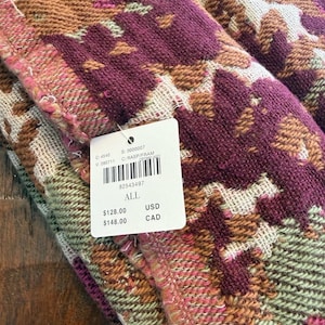 May include: Close-up of a folded textile with a geometric pattern in shades of burgundy, brown, pink, and green. The fabric has a textured appearance. A white tag with a barcode and text is attached to the textile.