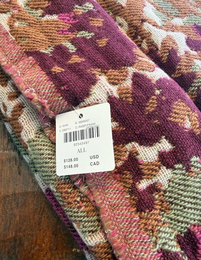 May include: Close-up of a folded textile with a geometric pattern in shades of burgundy, brown, pink, and green. The fabric has a textured appearance. A white tag with a barcode and text is attached to the textile.