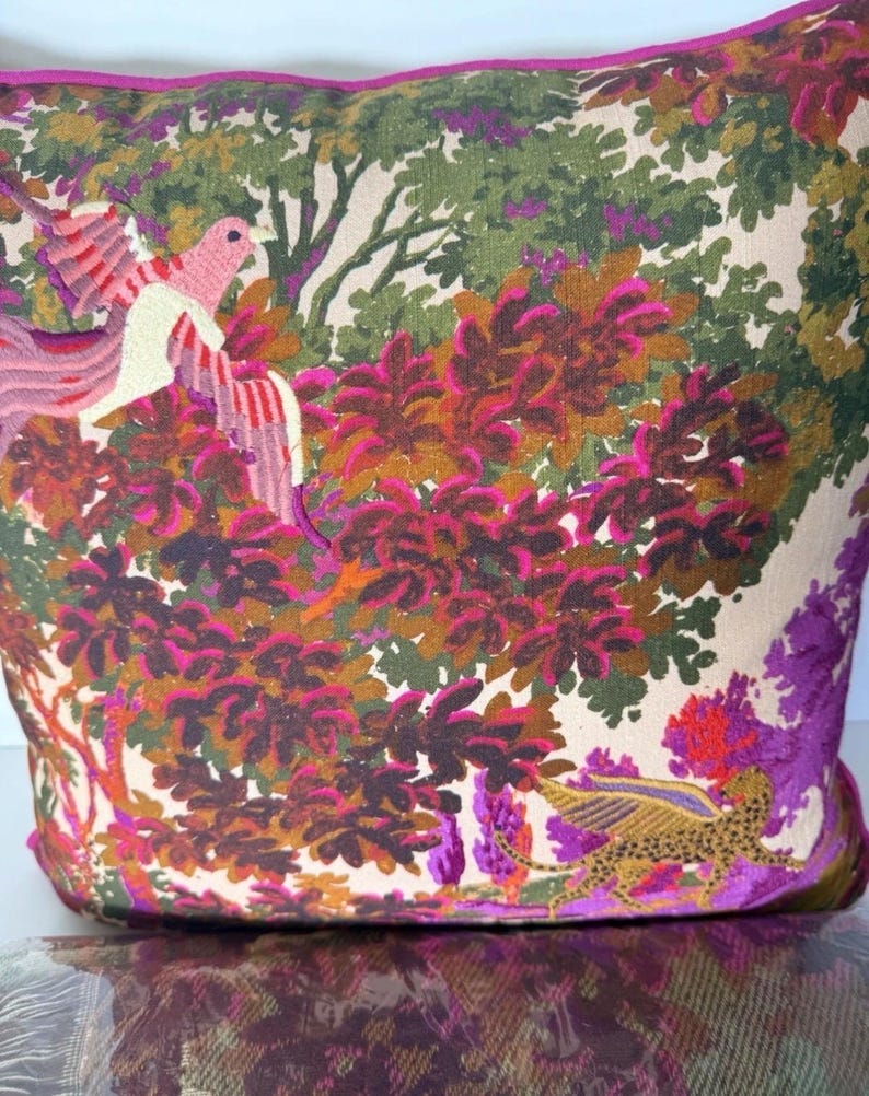 May include: A decorative pillow with a vibrant botanical design. The pillow features a colorful print of a bird, a leopard-like creature, and lush foliage in shades of pink, green, and purple. The pillow has a magenta border.