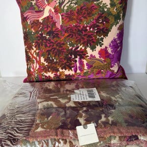 May include: A decorative pillow with a vibrant forest scene featuring birds and a leopard. The pillow has a pink border. Below is a folded blanket with a camouflage pattern in shades of brown, green, and pink.
