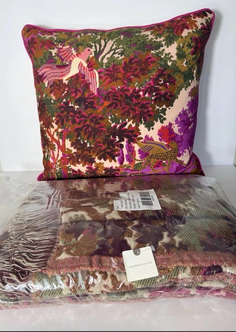 May include: A decorative pillow with a vibrant forest scene featuring birds and a leopard. The pillow has a pink border. Below is a folded blanket with a camouflage pattern in shades of brown, green, and pink.
