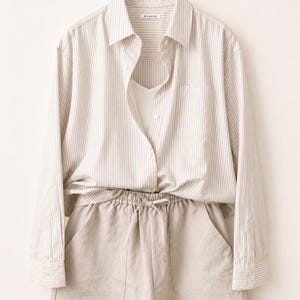 Puede incluir: Una camisa de manga larga abotonada con rayas verticales en beige y blanco, con cuello, un bolsillo en el pecho y una camiseta blanca debajo. Se combina con unos pantalones cortos beige con cintura el&aacute;stica de cord&oacute;n y dos bolsillos.