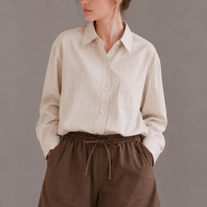 Puede incluir: Una camisa de manga larga con botones, un patr&oacute;n de rayas verticales en color crema y beige, cuello en punta y un bolsillo en el pecho, combinada con pantalones cortos de color marr&oacute;n oscuro liso con cintura el&aacute;stica y cord&oacute;n.