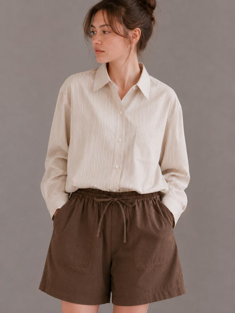 Puede incluir: Una camisa de manga larga con botones, un patr&oacute;n de rayas verticales en color crema y beige, cuello en punta y un bolsillo en el pecho, combinada con pantalones cortos de color marr&oacute;n oscuro liso con cintura el&aacute;stica y cord&oacute;n.