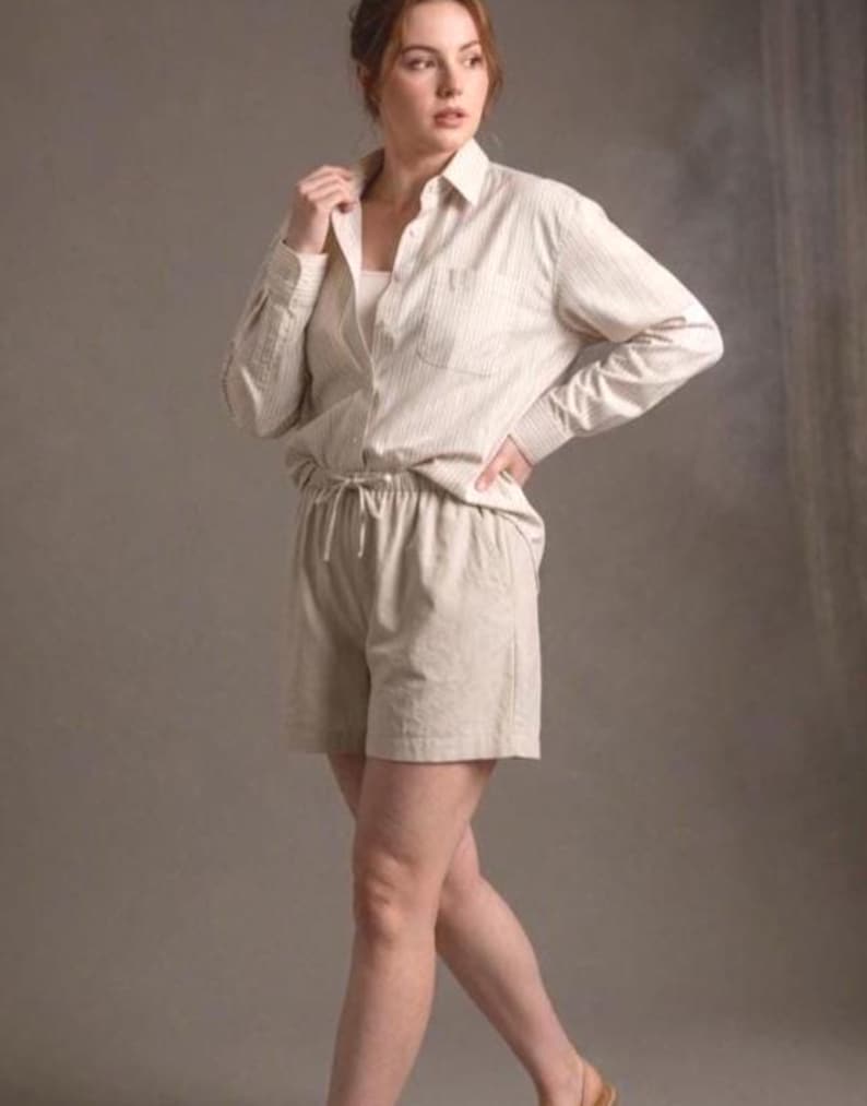 Puede incluir: Una camisa de manga larga abotonada en color beige claro con finas rayas verticales, que presenta un cuello y un bolsillo en el pecho. La camisa se combina con unos pantalones cortos beige a juego con cintura el&aacute;stica y cord&oacute;n.