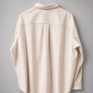 Puede incluir: Una camisa de manga larga con botones en color beige claro con finas rayas verticales marrones. La camisa presenta un cuello cl&aacute;sico, un canes&uacute; trasero con un pliegue central y pu&ntilde;os abotonados, mostrando la vista posterior.