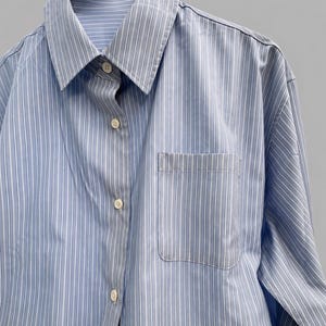 Puede incluir: Una camisa de manga larga con botones y un patr&oacute;n de rayas verticales en azul claro y blanco. La camisa presenta un cuello puntiagudo cl&aacute;sico, una tapeta frontal con botones de color claro y un bolsillo de parche en el pecho izquierdo.