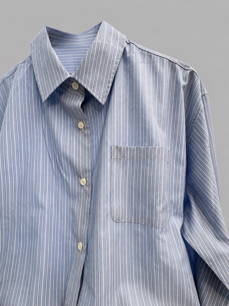 Puede incluir: Una camisa de manga larga con botones y un patr&oacute;n de rayas verticales en azul claro y blanco. La camisa presenta un cuello puntiagudo cl&aacute;sico, una tapeta frontal con botones de color claro y un bolsillo de parche en el pecho izquierdo.