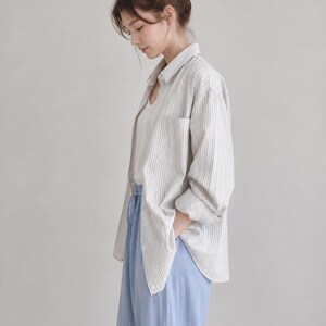 Puede incluir: Una camisa de manga larga abotonada con rayas verticales blancas y beige, sobre una camiseta interior blanca. El conjunto se completa con unos pantalones azul claro que tienen una cintura con cord&oacute;n y un corte holgado y relajado.