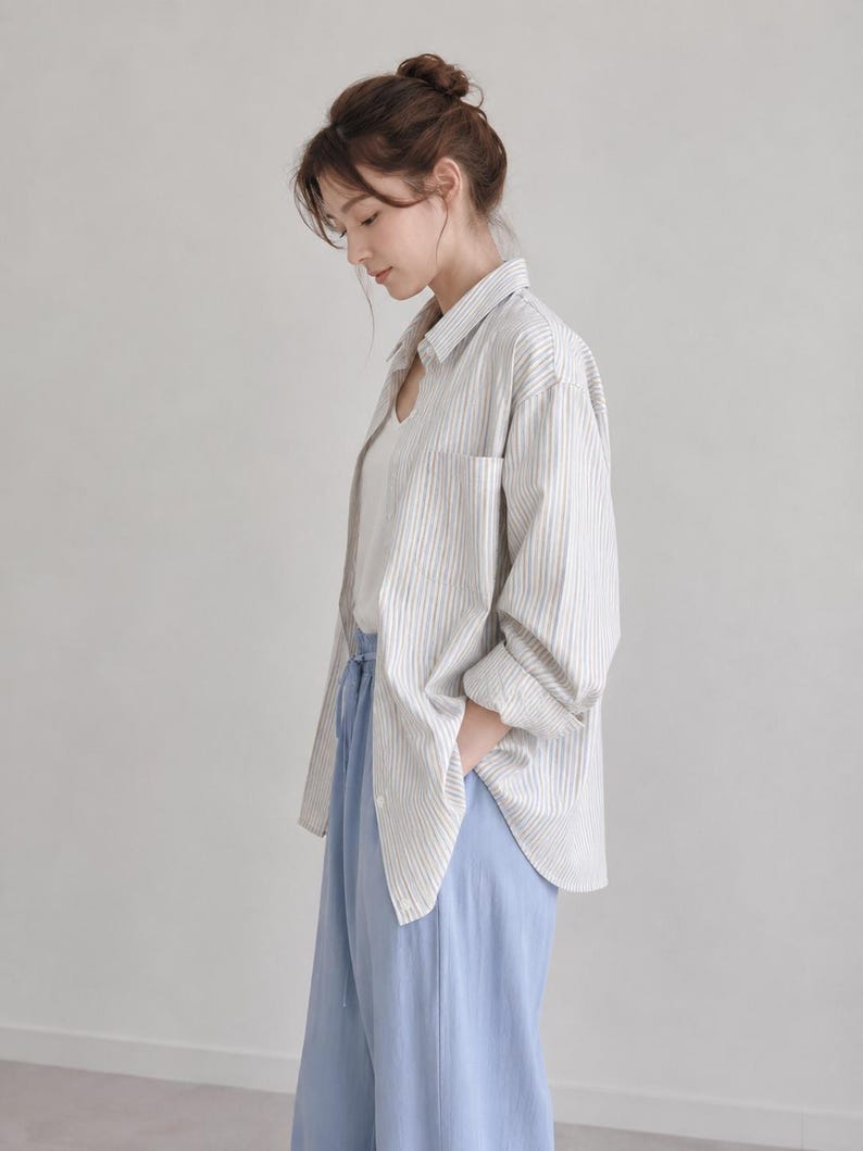 Puede incluir: Una camisa de manga larga abotonada con rayas verticales blancas y beige, sobre una camiseta interior blanca. El conjunto se completa con unos pantalones azul claro que tienen una cintura con cord&oacute;n y un corte holgado y relajado.