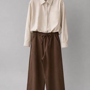 Puede incluir: Una camisa de manga larga abotonada con un patr&oacute;n de rayas verticales en beige y blanco, con cuello, un bolsillo en el pecho y pu&ntilde;os abotonados, combinada con pantalones anchos de color marr&oacute;n oscuro con cintura el&aacute;stica y cord&oacute;n.