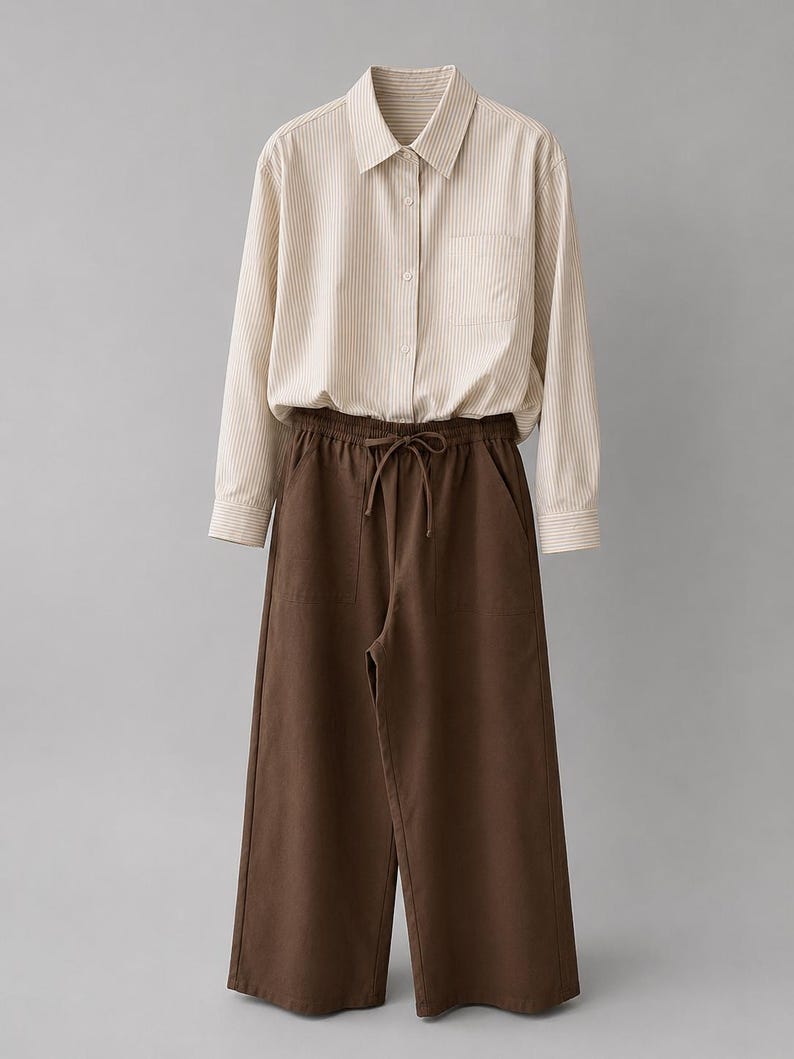 Puede incluir: Una camisa de manga larga abotonada con un patr&oacute;n de rayas verticales en beige y blanco, con cuello, un bolsillo en el pecho y pu&ntilde;os abotonados, combinada con pantalones anchos de color marr&oacute;n oscuro con cintura el&aacute;stica y cord&oacute;n.