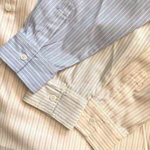 Puede incluir: Tres camisas de manga larga con botones superpuestas. Una camisa presenta rayas verticales en azul claro y blanco, mientras que las otras dos muestran rayas verticales en color tostado y blanco. Cada manga termina en un pu&ntilde;o abotonado.