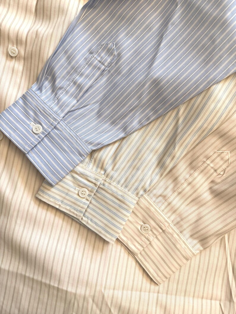 Puede incluir: Tres camisas de manga larga con botones superpuestas. Una camisa presenta rayas verticales en azul claro y blanco, mientras que las otras dos muestran rayas verticales en color tostado y blanco. Cada manga termina en un pu&ntilde;o abotonado.