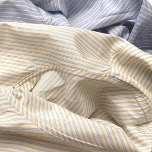 Puede incluir: Una camisa de manga larga con botones que presenta dos patrones de rayas distintos. La secci&oacute;n superior muestra rayas verticales en azul claro y blanco, mientras que la secci&oacute;n inferior exhibe un patr&oacute;n de rayas color canela y azul claro sobre un fondo crema.