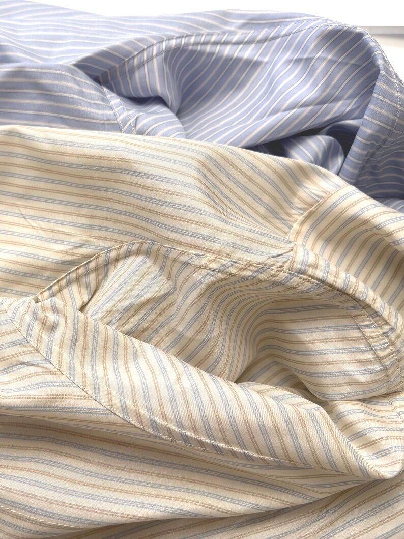 Puede incluir: Una camisa de manga larga con botones que presenta dos patrones de rayas distintos. La secci&oacute;n superior muestra rayas verticales en azul claro y blanco, mientras que la secci&oacute;n inferior exhibe un patr&oacute;n de rayas color canela y azul claro sobre un fondo crema.