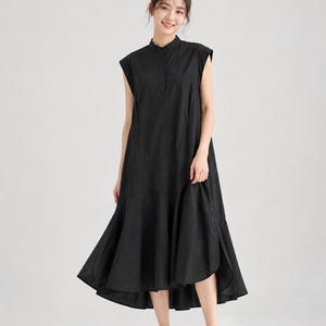 Pode incluir: Um vestido midi preto sem mangas com gola mandarim, uma carcela de botões parcial na frente e uma bainha assimétrica e rodada. O vestido tem uma silhueta larga e descontraída com cavas largas e um acabamento em tecido de cor sólida.