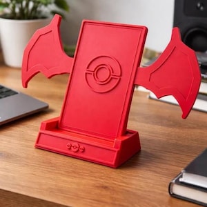 May include: A bright red phone stand with wing-shaped accents. The stand has a central panel with a circular emblem and a detailed base. It is designed to hold a phone or tablet, and is made of a solid material.
