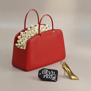 Könnte beinhalten: Eine rote Handtasche gefüllt mit Popcorn, mit zwei Henkeln. Ein goldener High-Heel-Schuh und ein schwarzes Etikett mit dem Text "THE DEVIL WEARS PRADA" liegen neben der Tasche. Ein kreatives Design.