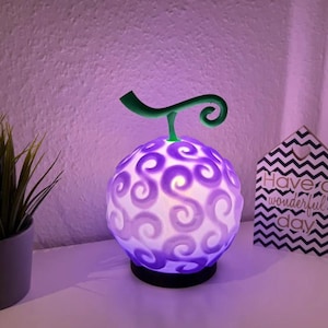 Puede incluir: Una lámpara decorativa morada brillante con forma de fruta y un tallo verde. La lámpara tiene un patrón de remolinos y se asienta sobre una base negra. También se ven una pequeña planta en maceta y un letrero con el texto "Have a wonderful day".