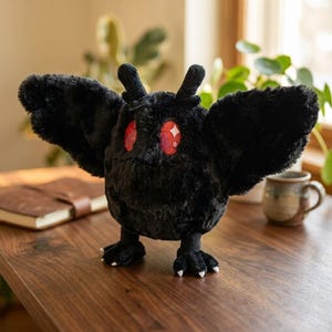 Pu&ograve; includere: Un peluche nero a forma di falena, con grandi ali spiegate e occhi rossi. Il peluche ha piccole antenne e zampe artigliate. &Egrave; posizionato su una superficie di legno.