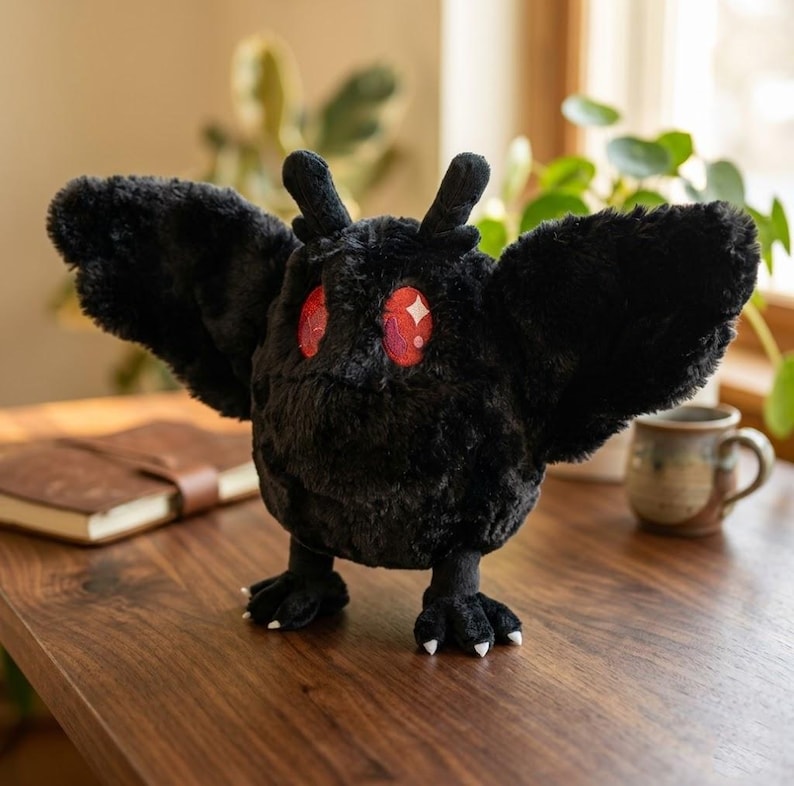 Pu&ograve; includere: Un peluche nero a forma di falena, con grandi ali spiegate e occhi rossi. Il peluche ha piccole antenne e zampe artigliate. &Egrave; posizionato su una superficie di legno.