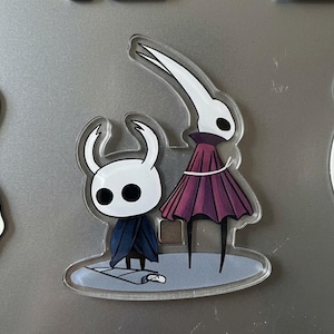 Puede incluir: Un imán acrílico transparente con dos personajes de dibujos animados. Uno es una figura pequeña con cabeza blanca y capa azul oscuro. El otro es una figura alta con cabeza blanca que lleva un vestido morado oscuro. El imán tiene un fondo gris.