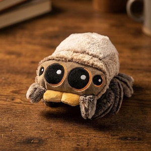 May include: A plush toy spider with a fuzzy, light brown body and a darker brown head. The spider has large, black eyes with white highlights and orange accents. Its legs are a darker shade of brown, and it has a yellow mouth.