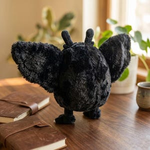 Pu&ograve; includere: Un peluche nero a forma di pipistrello con le ali spiegate, appoggiato su una superficie di legno. Il pipistrello ha una consistenza morbida e dettagli curati. Due quaderni rilegati in pelle marrone sono in primo piano.