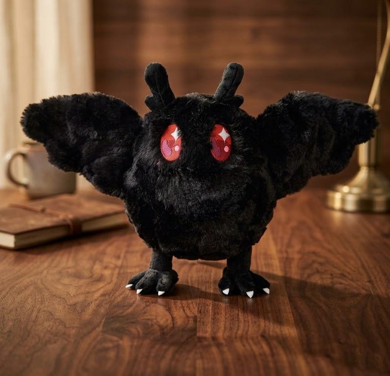 Pu&ograve; includere: Un peluche nero a forma di falena, con grandi occhi rossi espressivi e dettagli bianchi. Il giocattolo ha ali spiegate e piccoli piedi con artigli bianchi. La falena &egrave; posizionata su una superficie di legno.