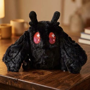 Pu&ograve; includere: Un peluche nero a forma di falena con grandi occhi rossi espressivi e una consistenza soffice. Il giocattolo ha grandi appendici simili ad ali e due piccole antenne. &Egrave; appoggiato su una superficie di legno.