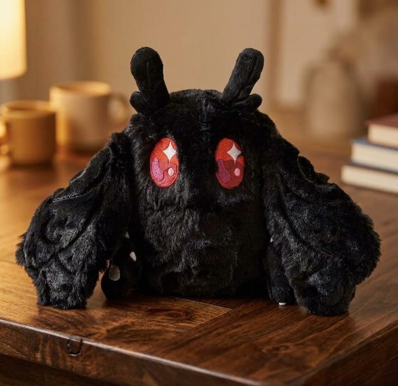 Pu&ograve; includere: Un peluche nero a forma di falena con grandi occhi rossi espressivi e una consistenza soffice. Il giocattolo ha grandi appendici simili ad ali e due piccole antenne. &Egrave; appoggiato su una superficie di legno.