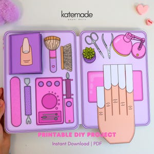 May include: A paper craft set featuring a purple case with illustrations of nail care tools including a brush, file, cuticle pusher, lamp, and scissors. A paper hand with white nail tips is attached to the right side. The text reads katemade paper dolls.