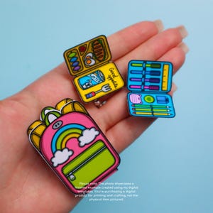 May include: A collection of colorful, cartoon-style pins featuring a pink backpack with a rainbow, a yellow lunchbox with food, and a blue pencil case. The lunchbox has the words "Good appetite" and a water bottle illustration. The pins are held in a hand.