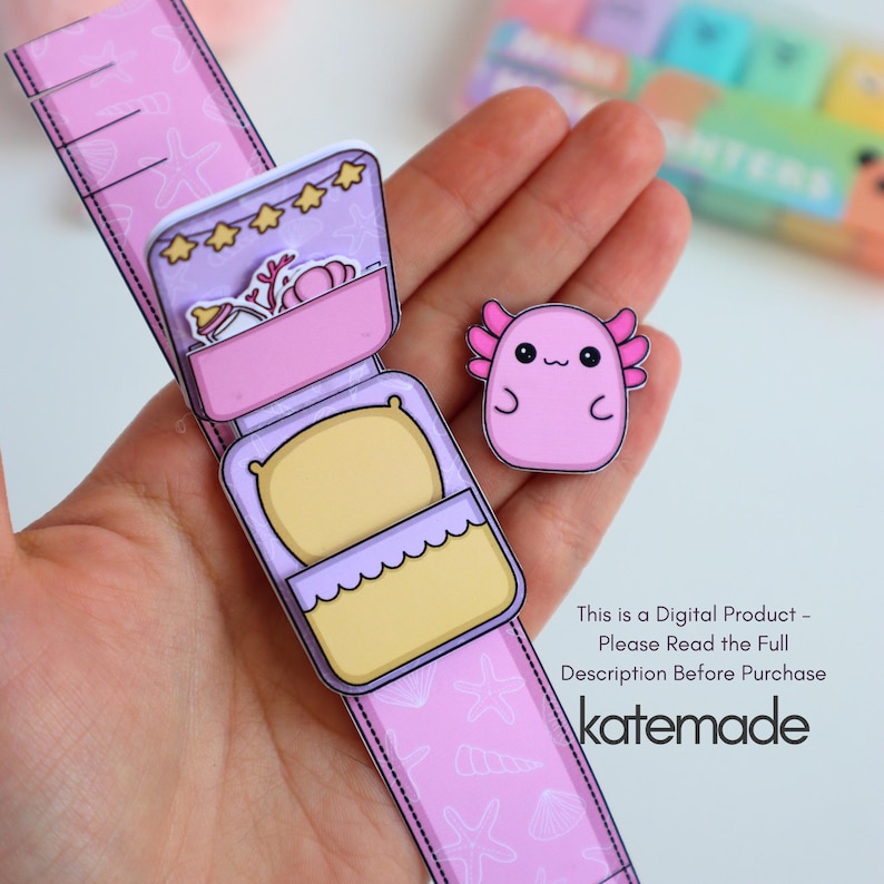 May include: A paper craft featuring a pink watch-like design with a bed and a small axolotl. The watch has a bed with a yellow pillow and a compartment with a bottle and stars. The axolotl is pink with a smiling face. The text "katemade" is visible.