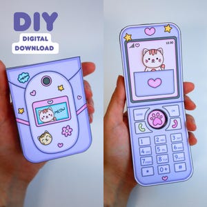 May include: A paper craft flip phone in light purple with cat-themed graphics, including a screen displaying a cartoon cat and the text MEOW. The device features a keypad with a paw print center button, star accents, and hearts. Text reads DIY DIGITAL DOWNLOAD.