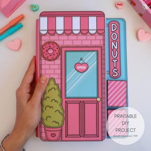 以下が含まれることがあります： ストライプのオーニング、ドーナツの看板、縦書きの「DONUTS」の文字が入った、ピンクと白のドーナツ屋さんのペーパークラフト。お店には、ハート型の「OPEN」の看板が付いたピンク色のドアがあります。手でクラフトを持っています。