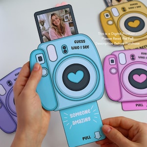 May include: A light blue paper camera with the text "Guess Who I See" and a heart-shaped lens. A photo of a woman is displayed on top. The camera has a pull-out tab that says "Someone Amazing" and "Pull". Other cameras in yellow and pink are also visible.