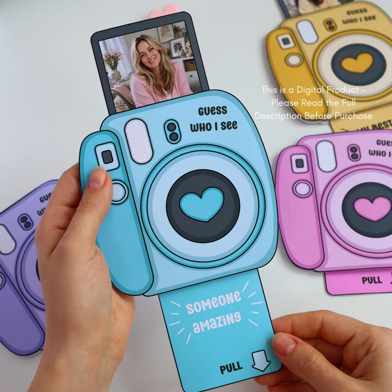 May include: A light blue paper camera with the text "Guess Who I See" and a heart-shaped lens. A photo of a woman is displayed on top. The camera has a pull-out tab that says "Someone Amazing" and "Pull". Other cameras in yellow and pink are also visible.