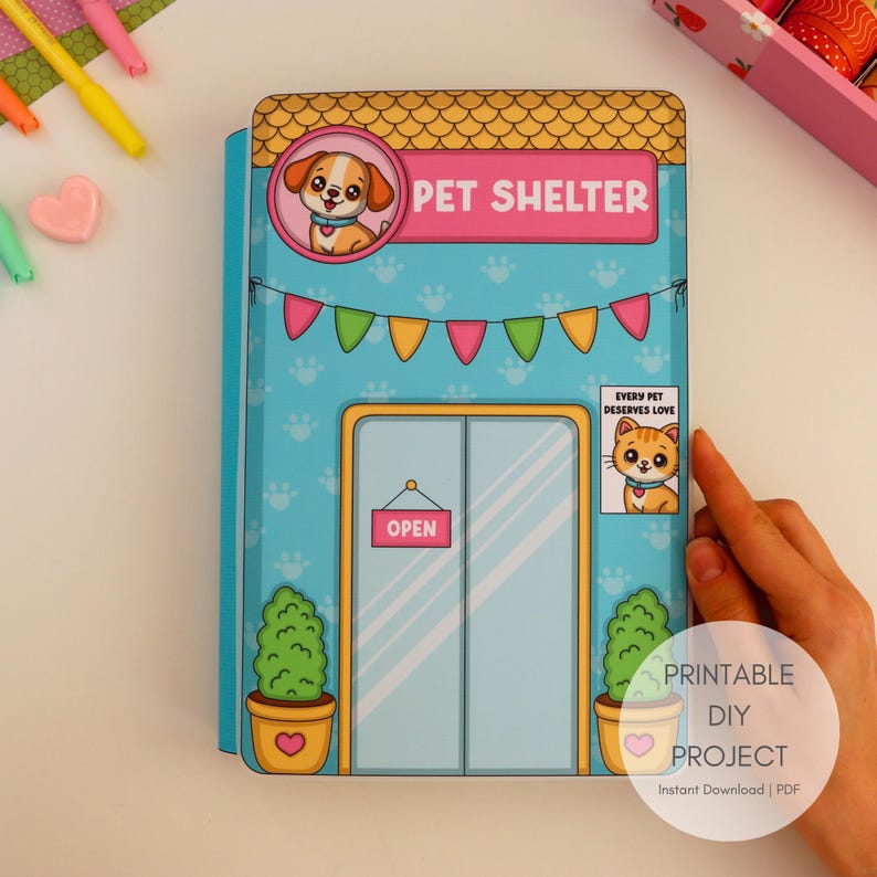 May include: A colorful DIY printable pet shelter project. The front of the shelter is light blue with paw prints and a sign that says "Pet Shelter". The shelter has a door with an "Open" sign and a poster that says "Every Pet Deserves Love".