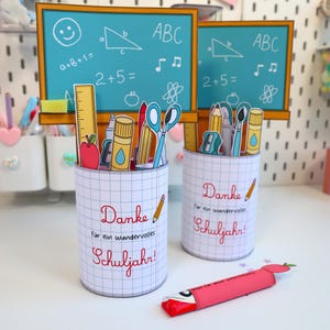以下が含まれることがあります： 学校をテーマにした装飾が施された2つのペン立て。各ホルダーはグリッドパターンで、「Danke für ein wundervolles Schuljahr！」という言葉が赤色で書かれています。数式や図が描かれたミニチュア黒板がホルダーの後ろにあります。ピンク色のキャンディーの包み紙が前景にあります。