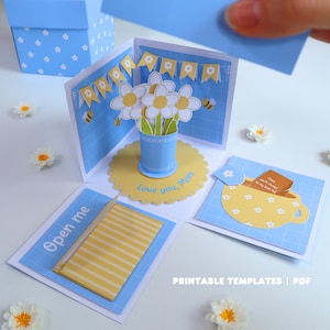 Könnte beinhalten: Eine handgefertigte blaue und weiße Papier-Explosionsbox mit floralem Thema. Die Box zeigt ein Pop-up-Blumenarrangement, eine Teetasse und den Text "Love you, Mom". Die Box ist mit Bienen und Bannern verziert.