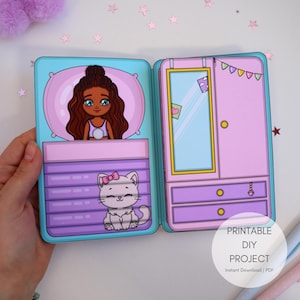 Könnte beinhalten: Ein druckbares DIY-Projekt mit einer Papierszene. Die Szene zeigt ein Mädchen im Bett, eine Katze und einen Kleiderschrank. Die Farbpalette ist pastellfarben mit Rosa-, Lila- und Blautönen. Der Text "PRINTABLE DIY PROJECT" ist sichtbar.