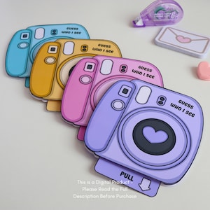 May include: A stack of colorful camera-shaped cards in blue, yellow, pink, and purple. Each card has a heart-shaped lens and the text "guess WHO I see". A small pink heart, a white envelope with a heart, and a purple tape dispenser are also visible.