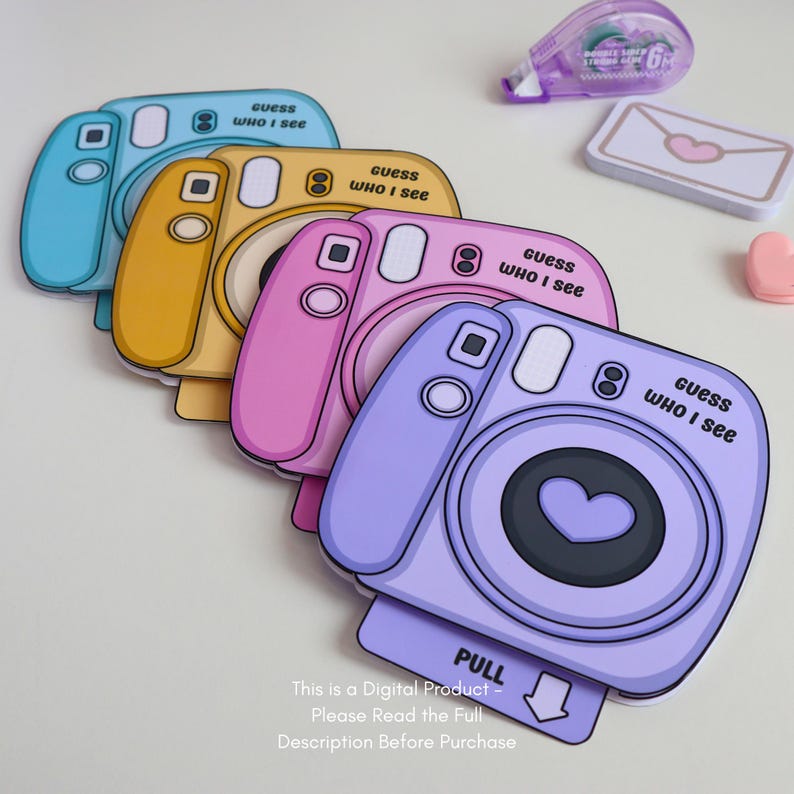 May include: A stack of colorful camera-shaped cards in blue, yellow, pink, and purple. Each card has a heart-shaped lens and the text "guess WHO I see". A small pink heart, a white envelope with a heart, and a purple tape dispenser are also visible.