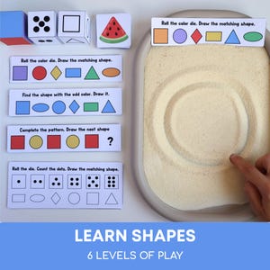 以下が含まれることがあります： ベージュの砂トレイ、色や形が描かれたサイコロ、そしてRoll the color die、Draw the matching shape、Find the shape with the odd color、Complete the patternなどのテキストが書かれた学習カードを含む感覚学習セット。